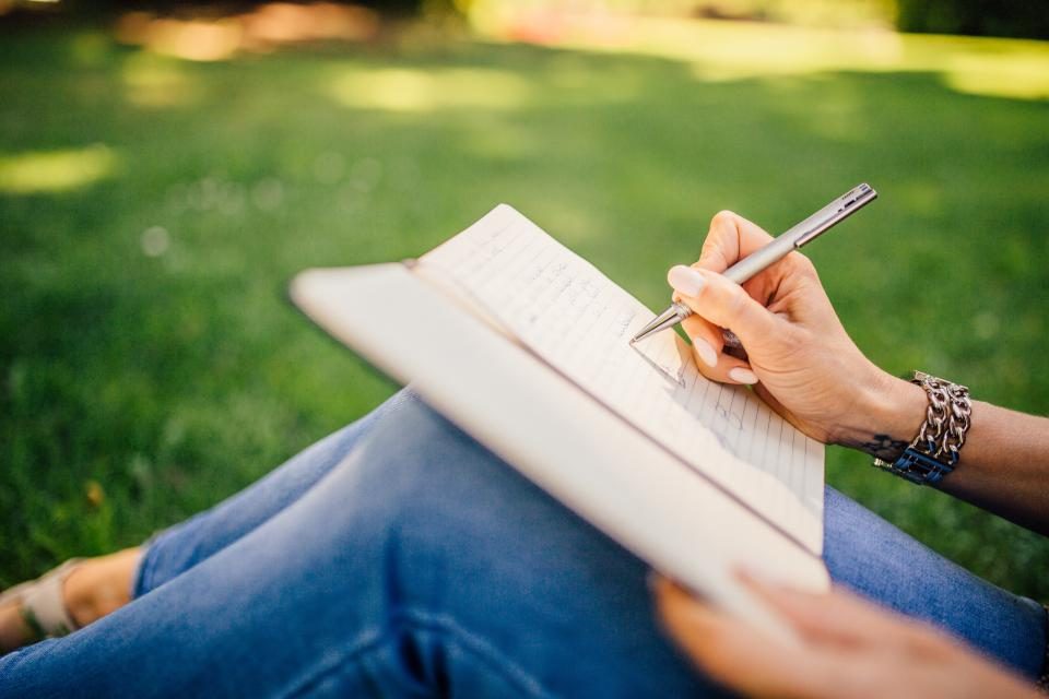 Unas manos escribiendo en un cuaderno apoyado sobre las rodillas, al aire libre.